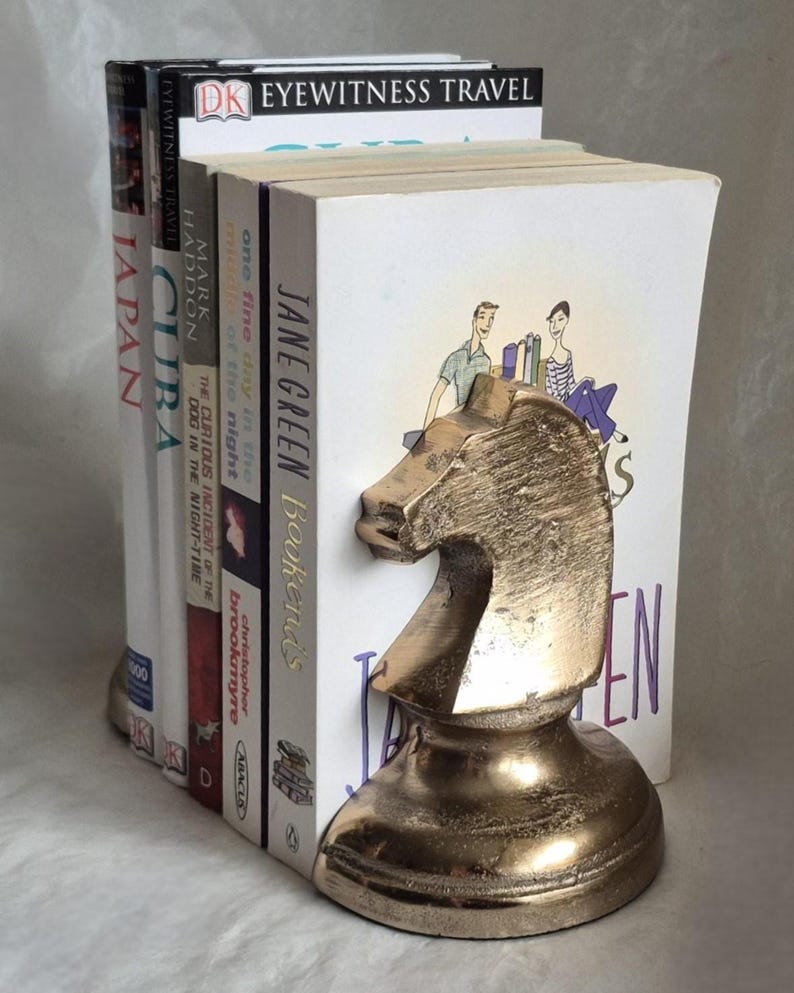 May include: A gold-colored metal knight chess piece bookend holding a stack of books. The books have titles like "Japan", "Cuba", and "Jane Green Bookends". The bookend is a decorative item for home or office.