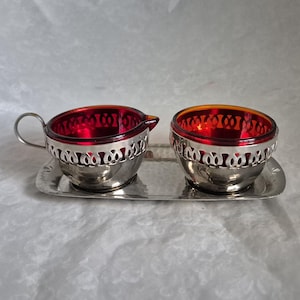 May include: A silver creamer and sugar bowl set with red glass inserts and a silver tray. The set has a decorative, ornate design around the rim of each bowl.