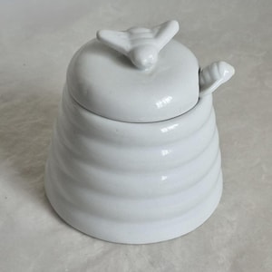Peut inclure: Pot à miel en céramique blanche avec couvercle. Le couvercle est orné d'une poignée en forme d'abeille, et le pot est conçu comme une ruche. Le pot est de couleur claire et neutre, avec une finition brillante. Article de cuisine.