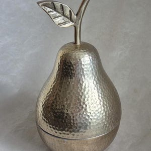 May include: A silver-colored pear-shaped container with a textured surface and a leaf and stem detail. The container is likely made of metal and has a lid that appears to be removable. The design is decorative.