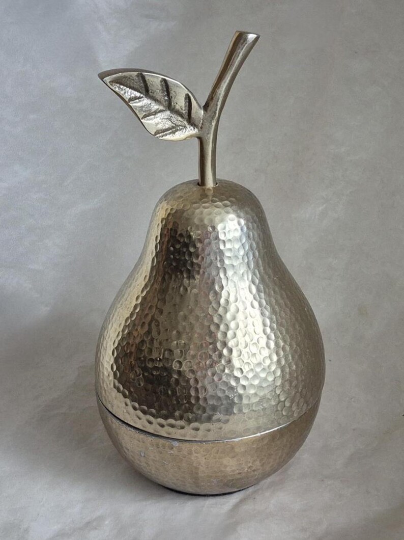 May include: A silver-colored pear-shaped container with a textured surface and a leaf and stem detail. The container is likely made of metal and has a lid that appears to be removable. The design is decorative.