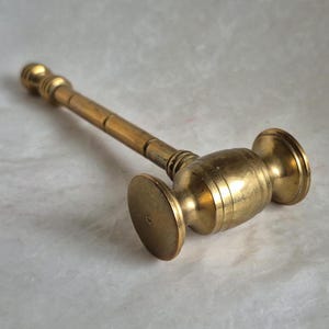 May include: A vintage brass gavel, featuring a cylindrical handle and a rounded head with a flat striking surface. The gavel is a warm gold color, with visible wear and tear, suggesting age and use. The design is simple and classic.
