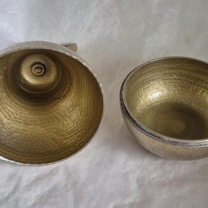 May include: Two metal bowls with a hammered texture. One is a large, open bowl with a handle, and the other is a smaller, round bowl. Both have a gold-colored interior and a silver-colored exterior.
