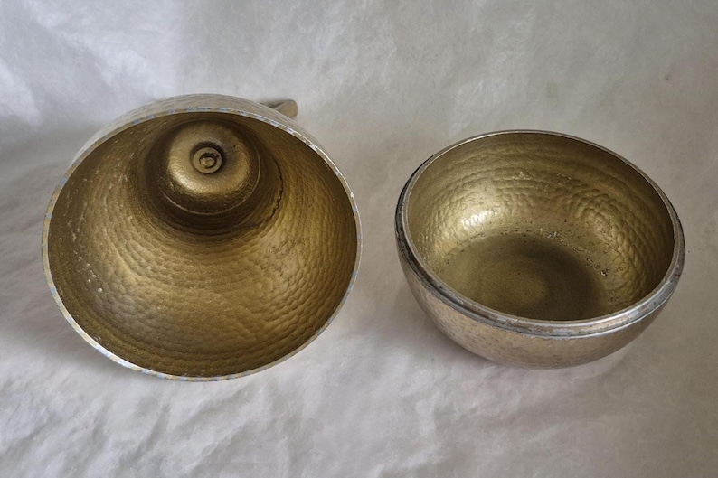 May include: Two metal bowls with a hammered texture. One is a large, open bowl with a handle, and the other is a smaller, round bowl. Both have a gold-colored interior and a silver-colored exterior.