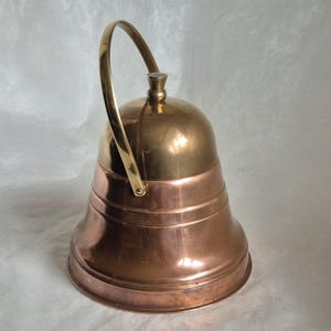 Può includere: Una campana in metallo lucido con una cupola superiore in ottone brillante e una base svasata color rame. La campana presenta un manico curvo in ottone fissato ai lati, un piccolo pomello sulla sommità e scanalature decorative orizzontali.