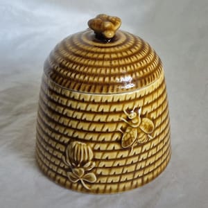 May include: A brown ceramic beehive-shaped jar with a lid and a bee-shaped handle. The jar has a textured design, with a bee and a flower relief on the side. The jar is likely for storing honey or other food items.