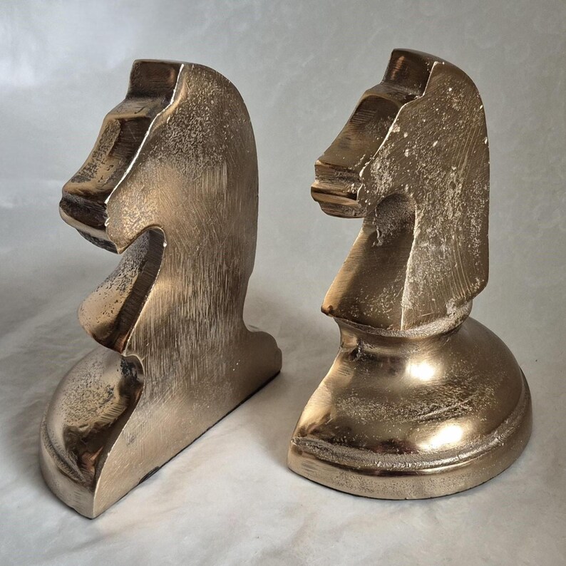 May include: Two bronze-colored knight chess piece bookends. The bookends are in the shape of a horse's head and neck. The bookends have a metallic finish and are displayed on a neutral background.