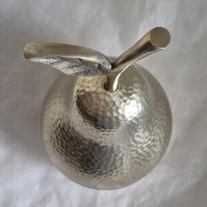 May include: A silver-colored apple-shaped decorative object with a textured surface and a stem and leaf detail. The apple has a hammered finish, giving it a unique, handcrafted appearance. The stem is cylindrical, and the leaf is detailed.