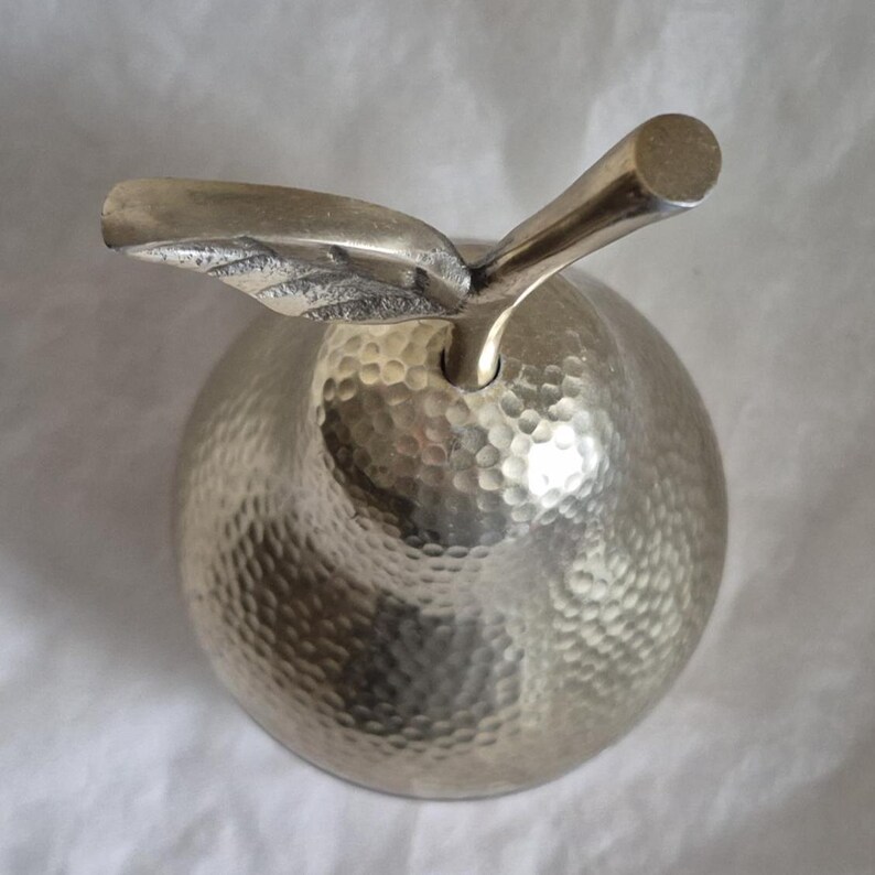 May include: A silver-colored apple-shaped decorative object with a textured surface and a stem and leaf detail. The apple has a hammered finish, giving it a unique, handcrafted appearance. The stem is cylindrical, and the leaf is detailed.
