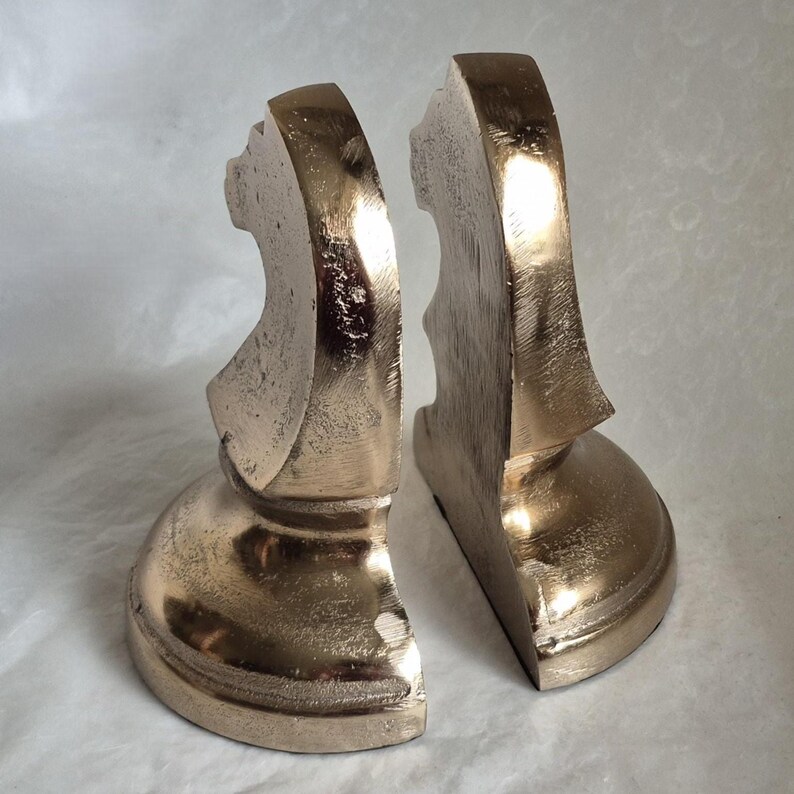 May include: A pair of metallic gold bookends. Each bookend has a decorative, curved design with a rounded base. The bookends are made of a solid material and are designed to hold books upright on a shelf or desk.