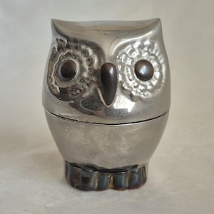 Könnte beinhalten: Eine silberfarbene Eulenfigur aus Metall mit glatter, reflektierender Oberfläche. Die Eule hat zwei dunkle, kreisförmige Augen und einen markanten dunklen Schnabel. Der Bereich um die Augen weist ein strukturiertes Muster auf, die Basis ist dunkel.
