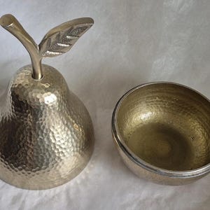 May include: A decorative metal pear-shaped container with a textured surface and a leaf-shaped handle. Beside it is a matching bowl, both with a hammered finish. The objects are likely for decorative or functional use.