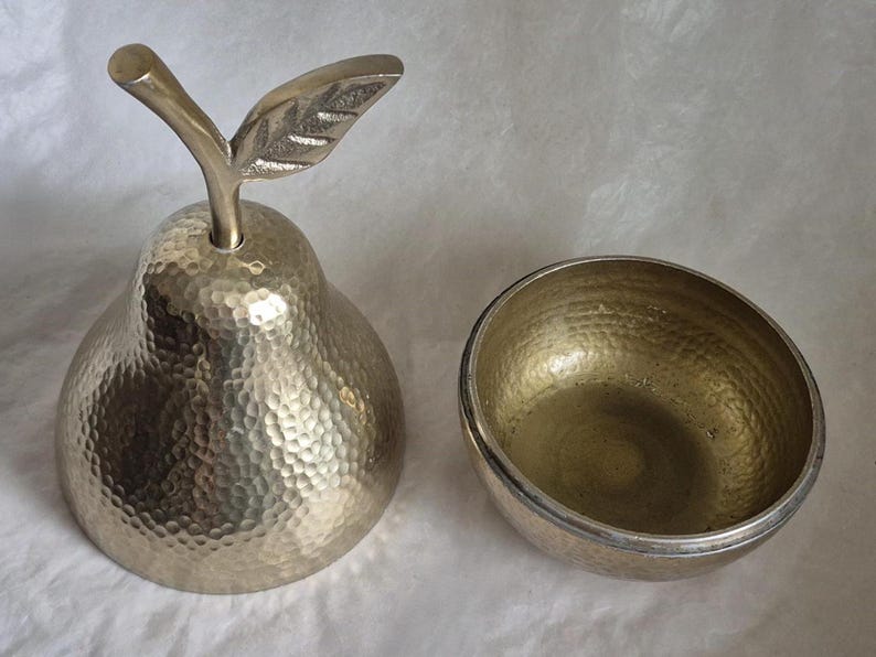 May include: A decorative metal pear-shaped container with a textured surface and a leaf-shaped handle. Beside it is a matching bowl, both with a hammered finish. The objects are likely for decorative or functional use.