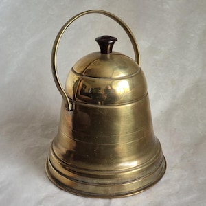 May include: A vintage brass bell-shaped container with a handle and a dark brown knob on top. The container has horizontal ridges and a shiny, reflective surface. The handle is arched and made of the same brass material.
