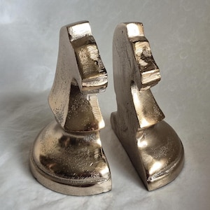 May include: A pair of metallic gold-colored bookends, each shaped like a chess knight. The bookends have a textured surface and are designed to stand upright, adding a decorative touch to any bookshelf or desk.
