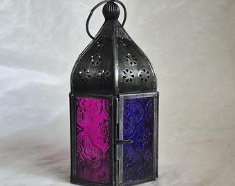 May include: A decorative metal lantern with a dark gray finish. The lantern features panels of patterned purple and blue glass. The top has decorative cut-out floral designs and a handle for hanging. The lantern is designed to hold a candle.