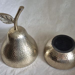 May include: A decorative pear-shaped container and a separate bowl, both with a hammered metal finish. The pear has a stem and leaf detail. The bowl has a black interior. The set is likely for home decor.