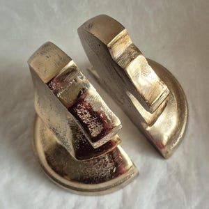 May include: Two brass-colored bookends, each with a geometric design. The bookends have a semi-circular base and a rectangular top, with a textured surface. The bookends are designed to hold books upright.