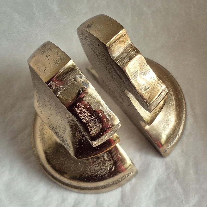 May include: Two brass-colored bookends, each with a geometric design. The bookends have a semi-circular base and a rectangular top, with a textured surface. The bookends are designed to hold books upright.