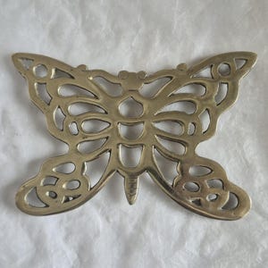 May include: A gold-toned metal trivet shaped like a butterfly with an intricate openwork design. The object features symmetrical cutouts throughout the wings and body, creating a decorative pattern with a smooth, polished metallic finish.