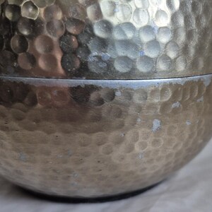 May include: Close-up of a silver-colored, hammered metal container. The surface has a textured, dimpled pattern. The container is in two parts, with a visible seam. The metal has some areas of discoloration.