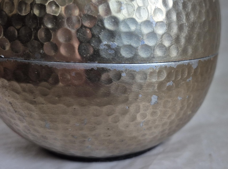 May include: Close-up of a silver-colored, hammered metal container. The surface has a textured, dimpled pattern. The container is in two parts, with a visible seam. The metal has some areas of discoloration.