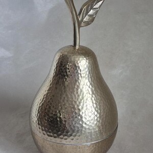 May include: A silver-colored pear-shaped decorative container with a textured surface. The container has a stem and leaf detail at the top. The container is likely made of metal and is designed for decorative purposes.