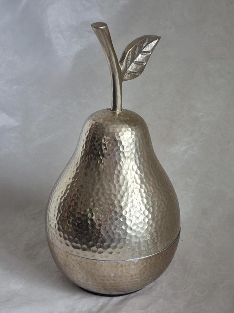May include: A silver-colored pear-shaped decorative container with a textured surface. The container has a stem and leaf detail at the top. The container is likely made of metal and is designed for decorative purposes.