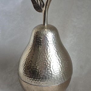 May include: Silver-colored pear-shaped decorative container with a textured surface and a stem with a leaf. The container is made of metal and has a lid. The pear is approximately 8 inches tall.