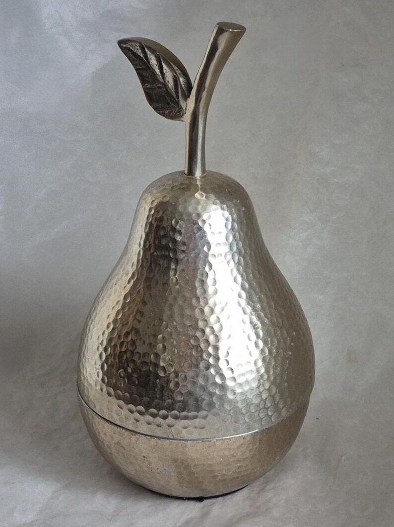 May include: Silver-colored pear-shaped decorative container with a textured surface and a stem with a leaf. The container is made of metal and has a lid. The pear is approximately 8 inches tall.
