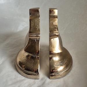 May include: A pair of metallic gold bookends. Each bookend has a rounded base and a vertical, rectangular support. The bookends are made of a shiny, textured metal, and are designed to hold books upright on a shelf.