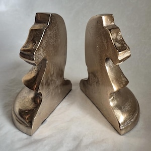 May include: Two bronze-colored bookends, each shaped like a Moai statue from Easter Island. The bookends have a weathered, metallic finish, with details of the face and head clearly visible. They are designed to stand upright and support books.