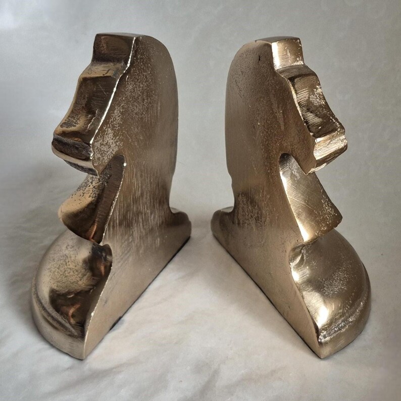 May include: Two bronze-colored bookends, each shaped like a Moai statue from Easter Island. The bookends have a weathered, metallic finish, with details of the face and head clearly visible. They are designed to stand upright and support books.