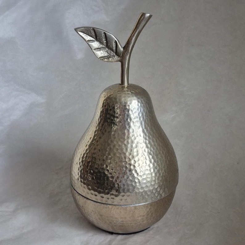 May include: Silver-colored pear-shaped decorative container with a textured surface. The container has a stem and leaf detail at the top. The pear-shaped container is likely used for storage or display.