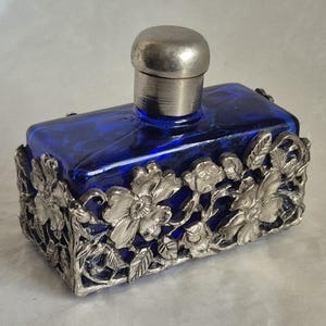 May include: A rectangular cobalt blue glass bottle featuring a silver-toned metal overlay with a detailed floral and leaf pattern. The bottle is topped with a smooth, cylindrical silver-toned metal cap that sits centred on the top surface.
