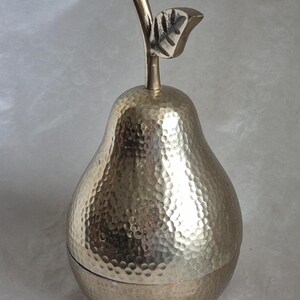 May include: A silver-colored pear-shaped container with a textured surface and a stem and leaf detail. The container is made of metal and has a lid. The pear-shaped container is a decorative item.