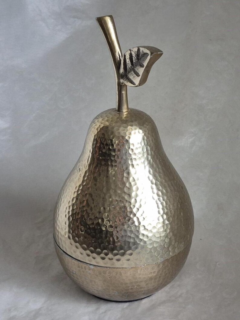 May include: A silver-colored pear-shaped container with a textured surface and a stem and leaf detail. The container is made of metal and has a lid. The pear-shaped container is a decorative item.
