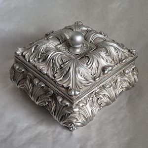 May include: A silver-coloured, square jewellery box with ornate, raised leaf and scroll detailing. The box has a small, round knob on the lid and sits on a decorative base. The overall design is classical and elegant.