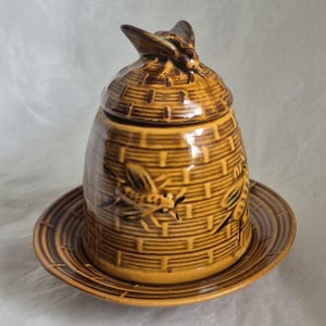 May include: A ceramic honey pot shaped like a traditional skep beehive with a matching saucer. The pot features a golden-brown glaze, a textured basket-weave pattern, and embossed bee motifs on the sides and the lid handle.