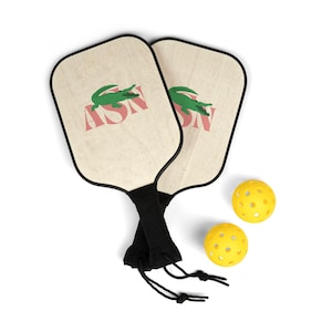 Könnte beinhalten: Zwei Pickleball-Schläger mit einem grünen Alligator-Logo und den Buchstaben "ASN" in Rot. Die Schläger haben schwarze Griffe und werden von zwei gelben, perforierten Pickleballs begleitet. Die Schläger sind aus Holz.