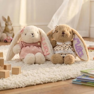 May include: Two plush bunny rabbits with long ears, wearing dresses, sit on a white rug. One bunny is cream-colored with a pink gingham dress, the other is brown with a polka-dot dress. The ears have names embroidered on them.