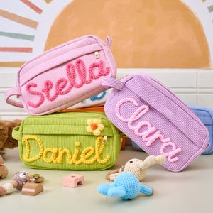 May include: A collection of corduroy cosmetic bags in various colors, including pink, green, and purple. Each bag features a name embroidered in a contrasting color, such as "Stella" in pink and "Daniel" in yellow. The bags have a zipper closure and a small handle.