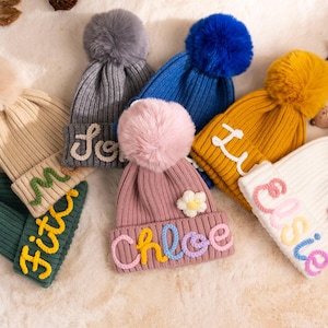 Puede incluir: Una colección de gorros de punto con pompones. Cada gorro presenta un nombre bordado en el borde, como "Chloe", "Ellie" y "Love". Los gorros son de varios colores, como rosa, amarillo y azul, ideales para el invierno.