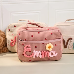 Könnte beinhalten: Rosa Cord-Mittagstasche mit Erdbeermuster und dem Namen "Emma" aus rosa und weißem Garn. Die Tasche hat einen Reißverschluss, eine kleine Vordertasche und eine gelbe Blumenapplikation. Weitere Taschen sind im Hintergrund zu sehen.