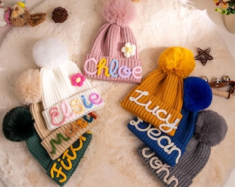 Puede incluir: Una colección de gorros de punto personalizados en varios colores, incluyendo rosa, blanco, amarillo, azul y gris. Cada gorro presenta un pompón esponjoso y un nombre bordado con hilo colorido. Algunos gorros tienen flores.