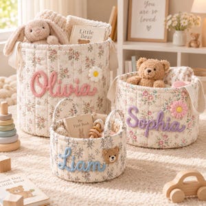Könnte beinhalten: Drei zylindrische Aufbewahrungskörbe aus Stoff mit cremefarbener Basis und Blumenmuster. Jeder Korb zeigt einen personalisierten Namen in erhabener Garnschrift: Olivia in Pink, Sophia in Lila und Liam in Blau, verziert mit Blumen- oder Bärenapplikationen.
