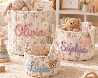Könnte beinhalten: Drei zylindrische Aufbewahrungskörbe aus Stoff mit cremefarbener Basis und Blumenmuster. Jeder Korb zeigt einen personalisierten Namen in erhabener Garnschrift: Olivia in Pink, Sophia in Lila und Liam in Blau, verziert mit Blumen- oder Bärenapplikationen.