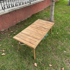 Puede incluir: Una mesa plegable de madera con un acabado natural. La mesa tiene un diseño sencillo con una tapa rectangular y cuatro patas. La mesa está sentada sobre una superficie cubierta de hierba.