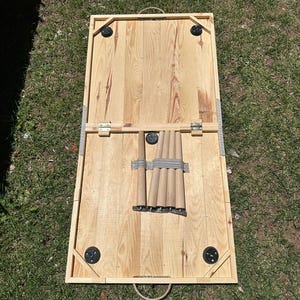 May include: A wooden cornhole board set, featuring a natural wood finish. The board has a rectangular frame with rounded corners and black corner supports. The legs are stored underneath. The board is designed for outdoor games.