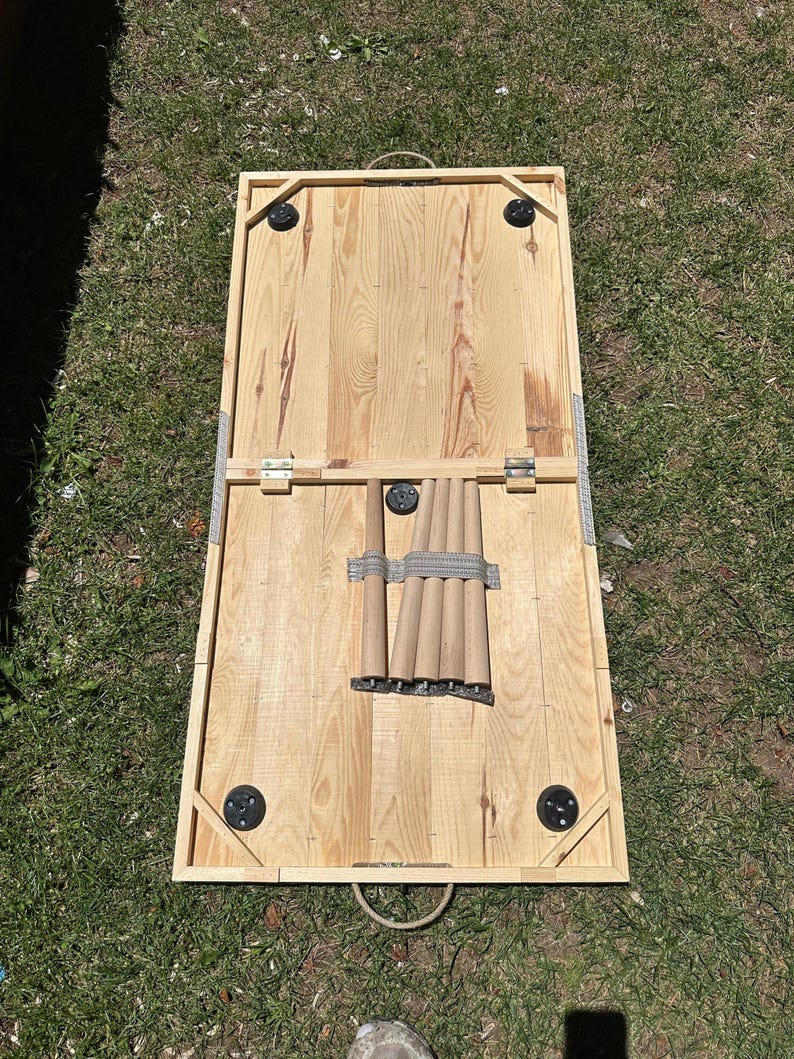 May include: A wooden cornhole board set, featuring a natural wood finish. The board has a rectangular frame with rounded corners and black corner supports. The legs are stored underneath. The board is designed for outdoor games.